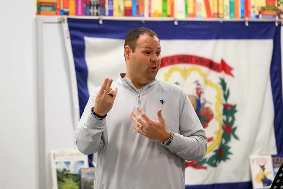 baker talks in front of the West Virginia flag at an elementary school