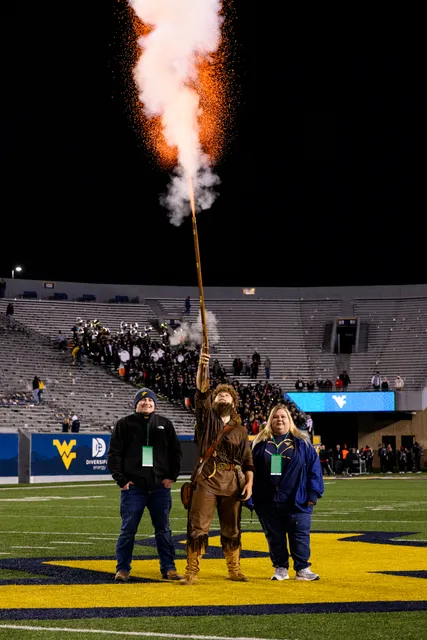 A muzzle flash appears above Mountaineer mascot Mikel Hager as he fires his rifle for a final time on Mountaineer Field. He's joined by his parents at his side.