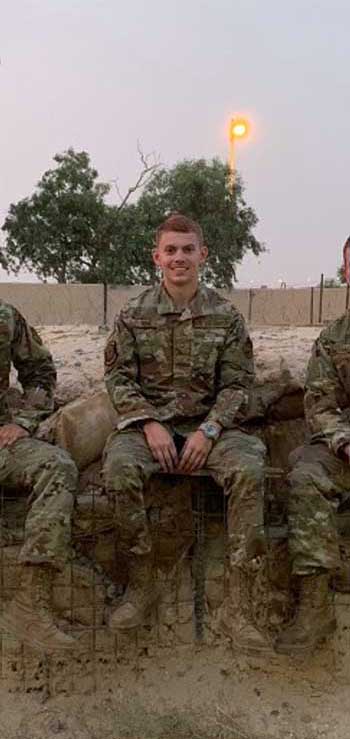 person in fatigues sits with others, desert sunset in background