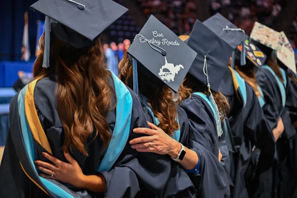 Graduates are seen from behind, arms around each other, singing 'Country Roads' while one cap reads 