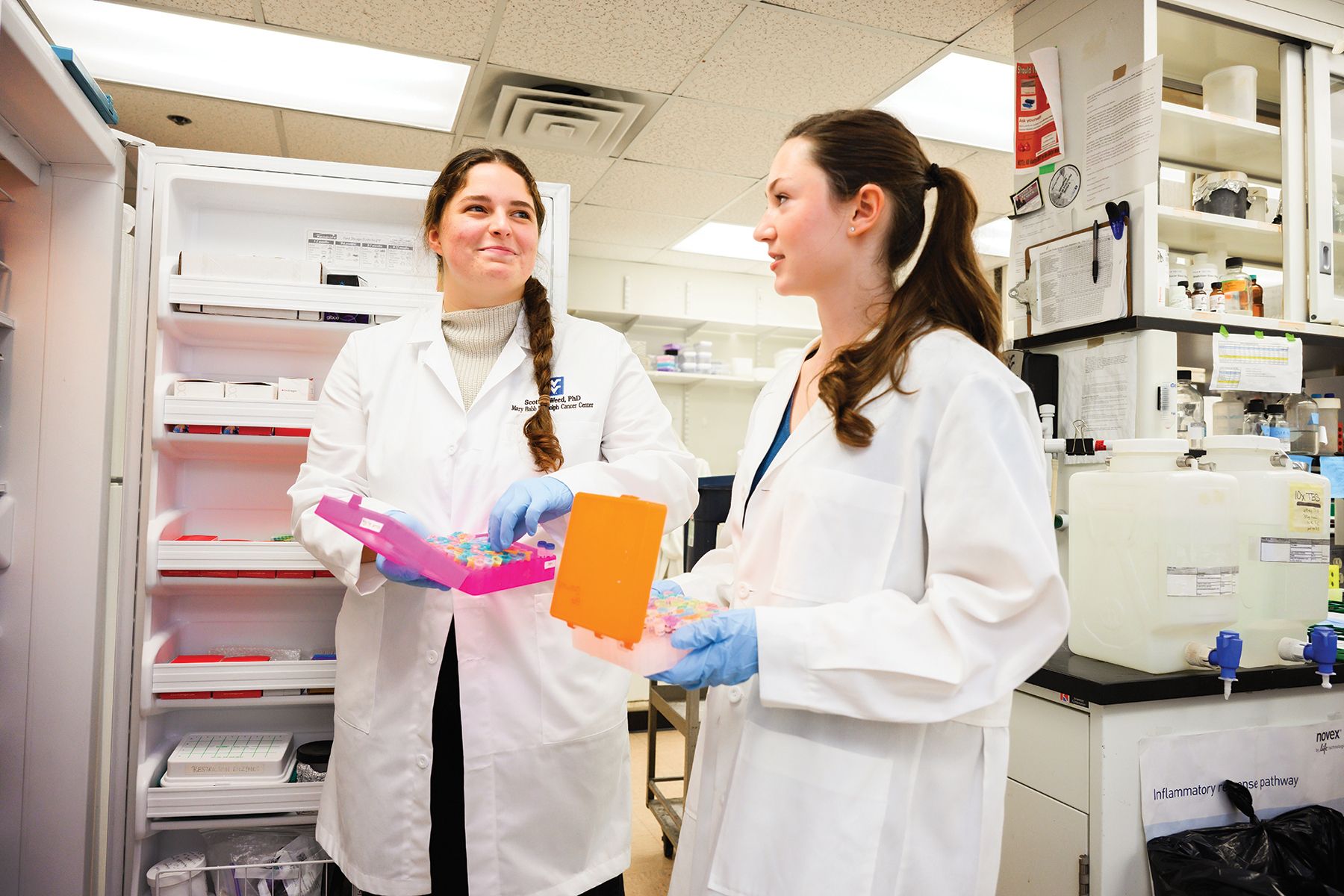 Two women in lab coats, look at each other, holding containers